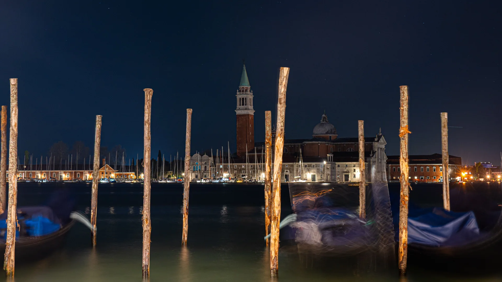 Una laguna oscura di notte, con paline d’ormeggio e il campanile di una chiesa sulla riva lontana, immagine rappresentativa del panorama nei pressi di San Giorgio Maggiore.